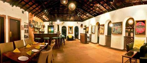 A restaurant with a long wooden table, wicker chairs, and a bar area with shelves of bottles.