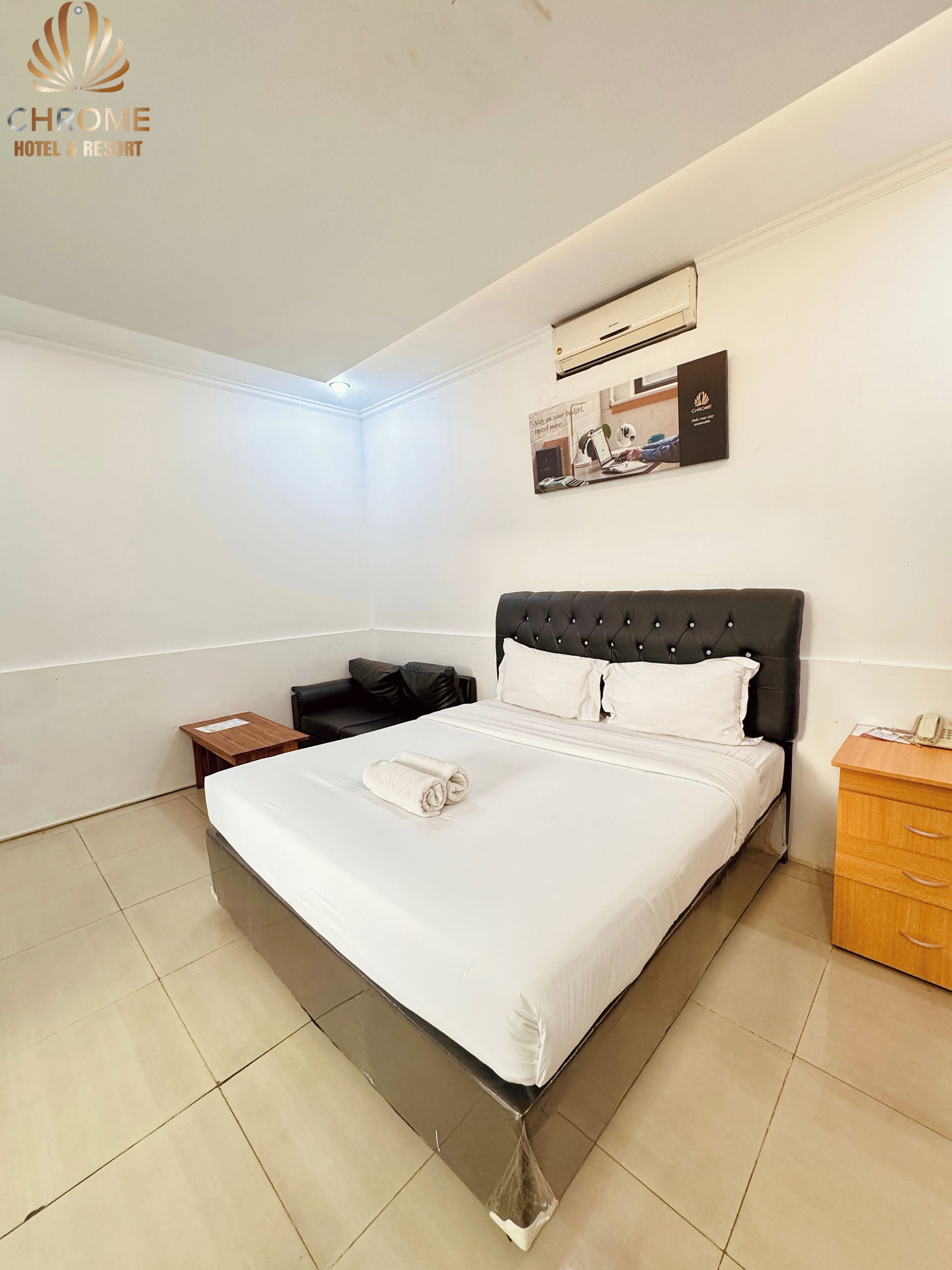 A hotel room with a large bed, a wooden nightstand, a black upholstered chair, a wooden dresser, and a wall-mounted air conditioner.