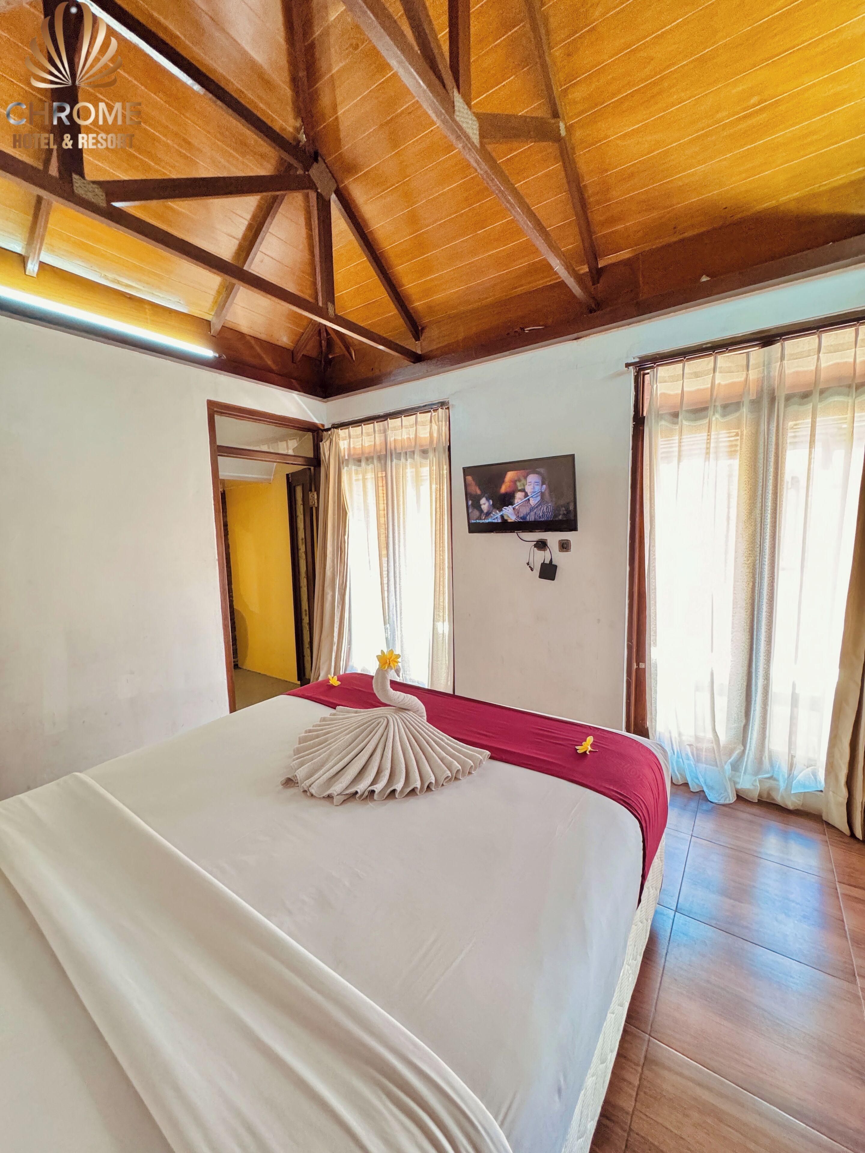 Ein Hotelzimmer mit Holzbalkendecke, einem Bett mit weißer Bettdecke und einem Zierhandtuch, einem Fernseher an der Wand und Vorhängen an den Fenstern.