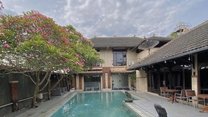 A swimming pool with a tiled deck, a covered seating area, and a building with a tiled roof in the background. - Chrome Hotel & Resort Solo (Colomadu)
