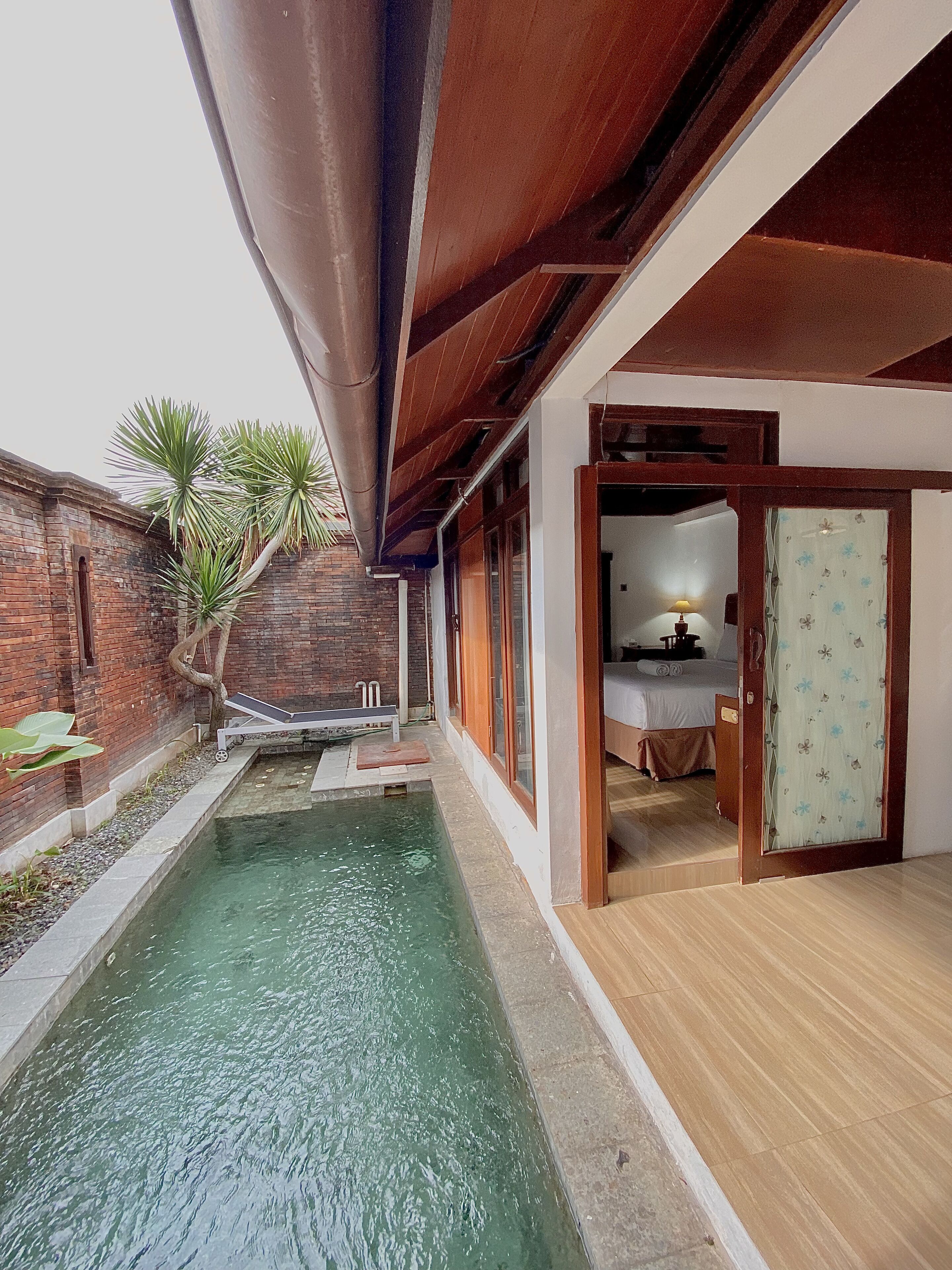 A pool area with a wooden deck, a brick wall, and a door leading to a bedroom.