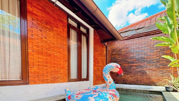 Montaje de comedor junto a la piscina, con un flotador de flamenco, una bandeja con platos de comida y un edificio de ladrillo al fondo.