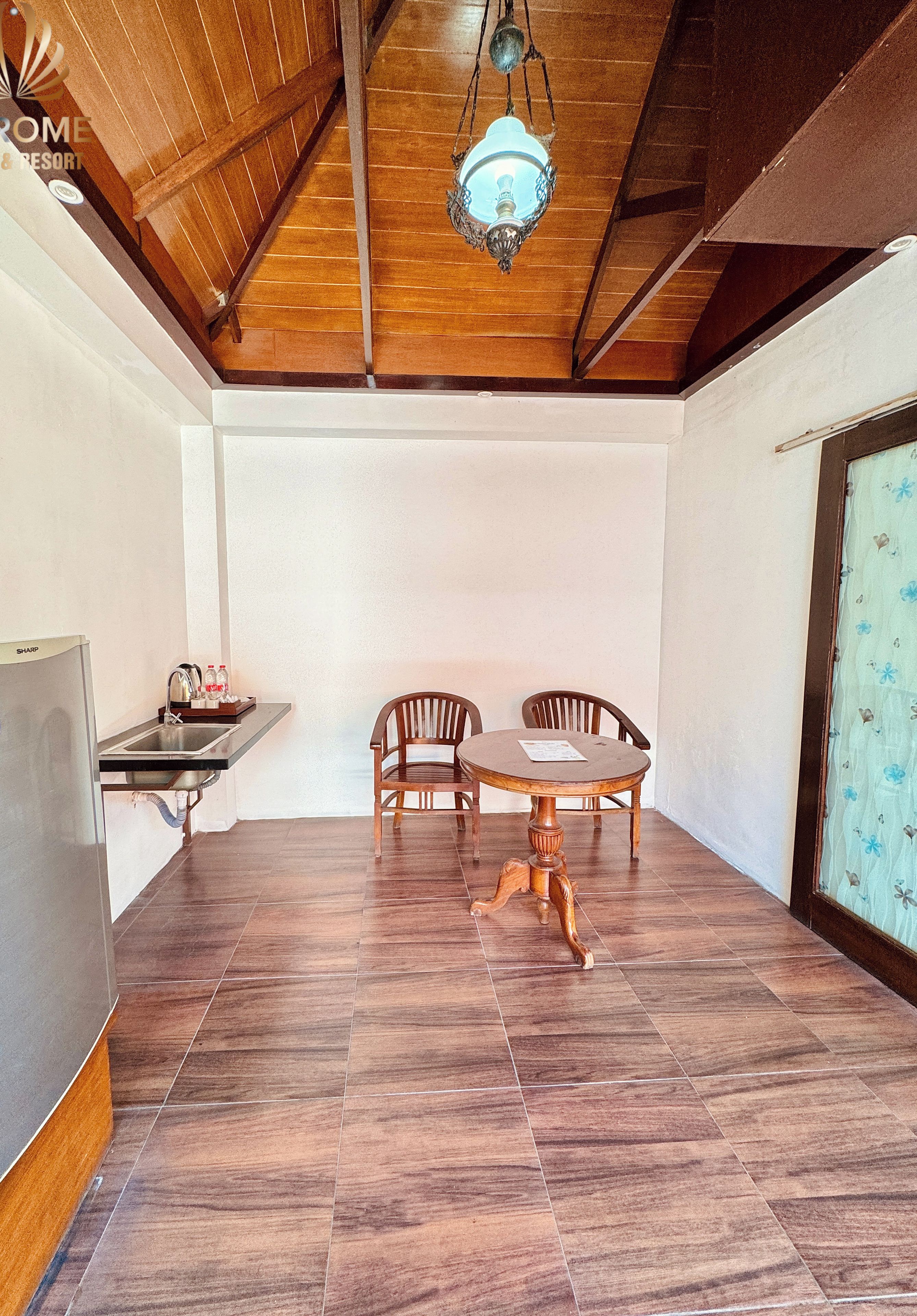 A dining area with a wooden table and chairs, a hanging light fixture, and a kitchen area with a refrigerator and sink.