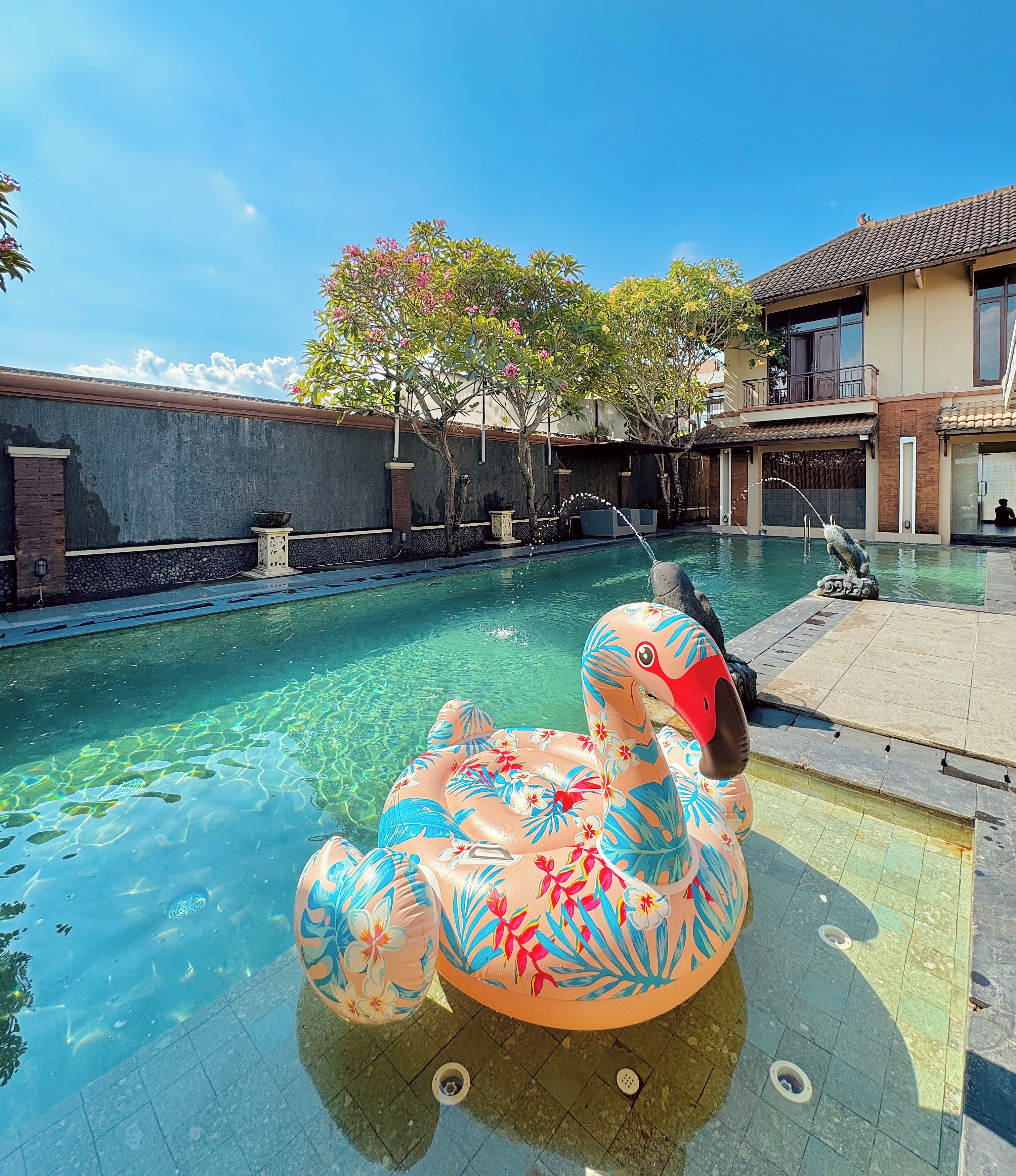 Una piscina con un flotador de flamenco decorativo, rodeada por un muro de ladrillo y árboles.