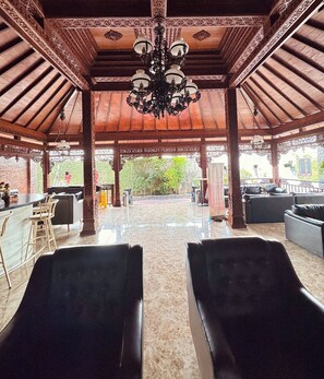 A spacious indoor area with a high ceiling, ornate wooden beams, and a central chandelier. There are several black cushioned seating areas and a bar with stools. - Chrome Hotel & Resort Solo (Colomadu)