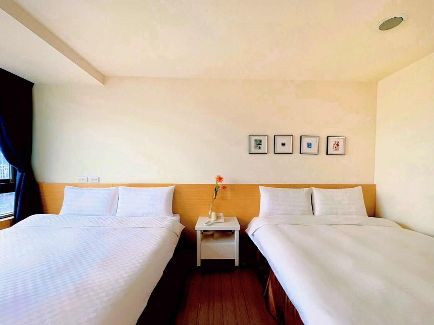A hotel room with two beds, a nightstand with a vase of flowers, and framed pictures on the wall.