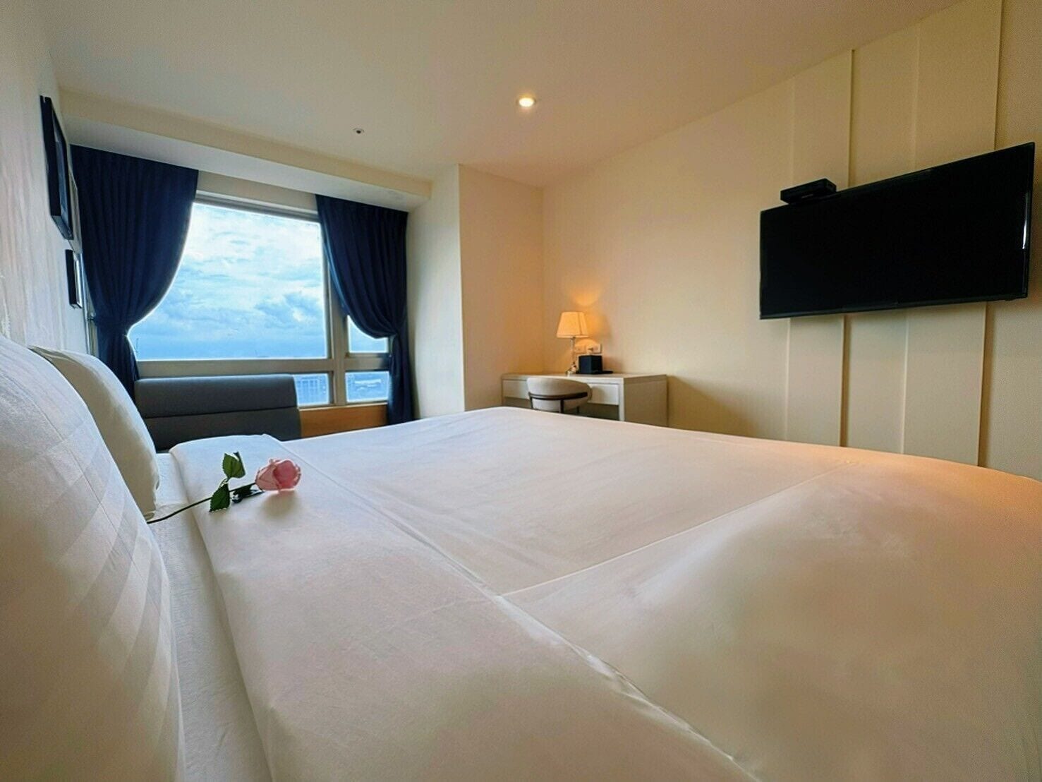 A hotel room with a large bed, a desk with a lamp, a TV mounted on the wall, and a window with curtains.