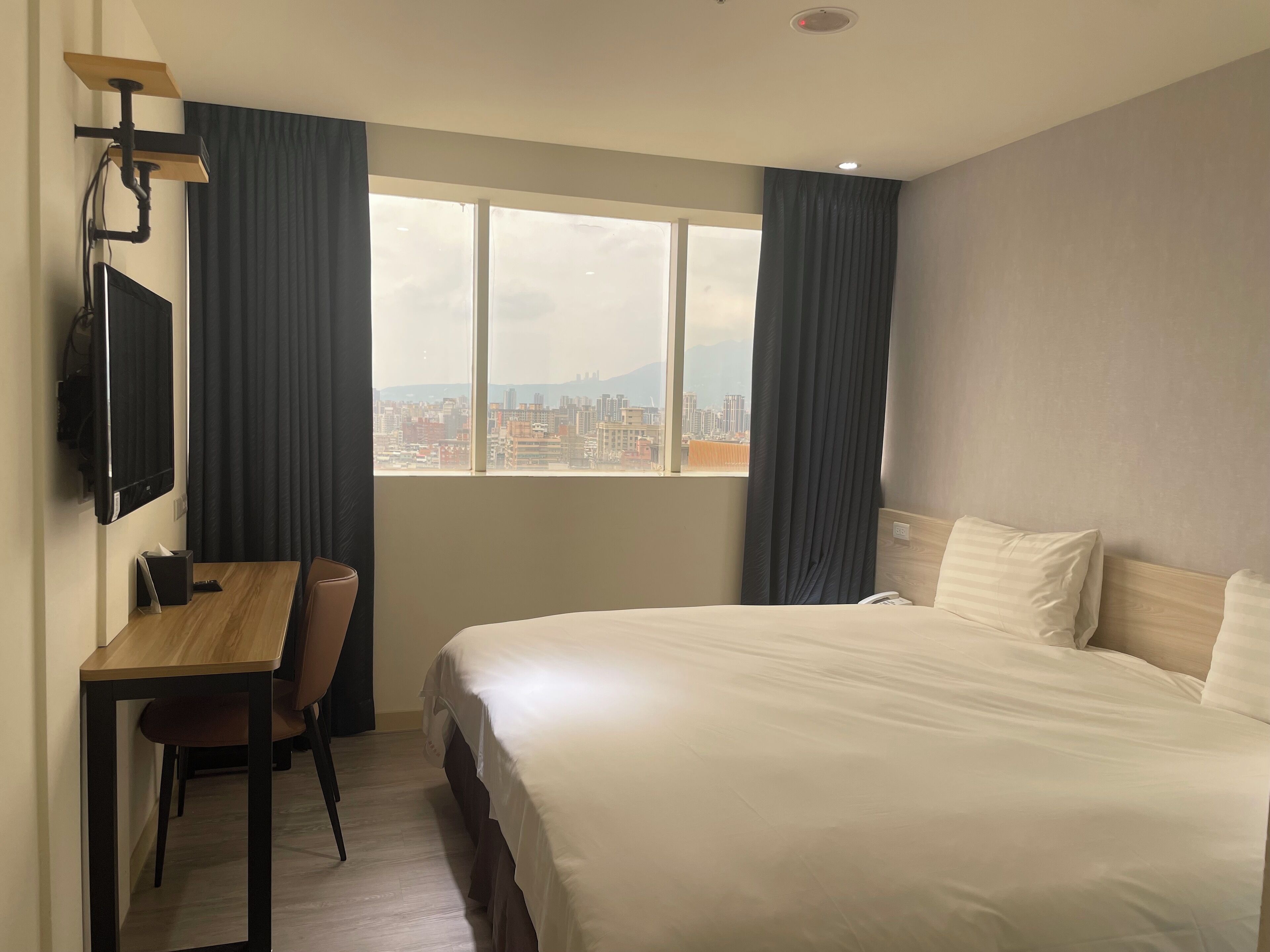 A hotel room with a bed, a desk, a chair, a TV, and a window with a city view.