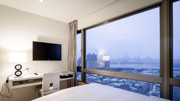 A hotel room with a bed, a desk with a computer, a TV, and a large window offering a city view. - We Meet Taipei Hotel (New Taipei City)