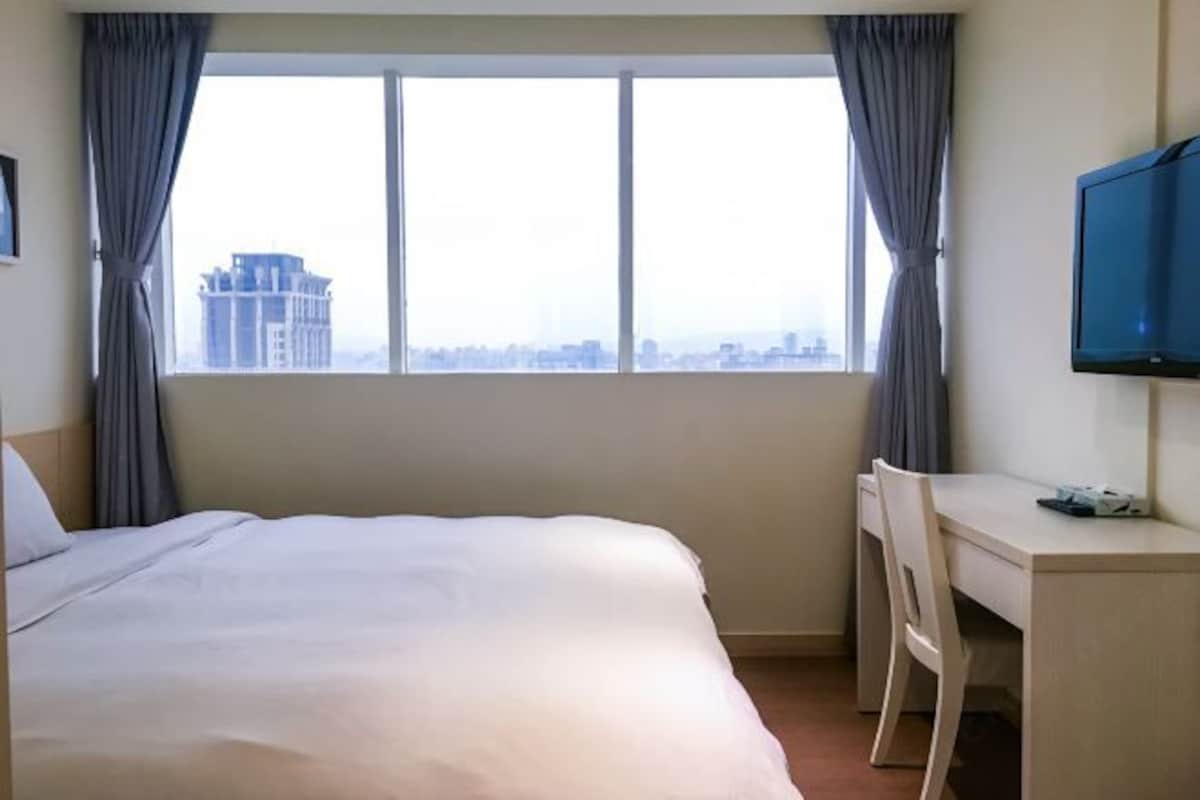A hotel room with a bed, a desk, a chair, a TV, and a large window with curtains.