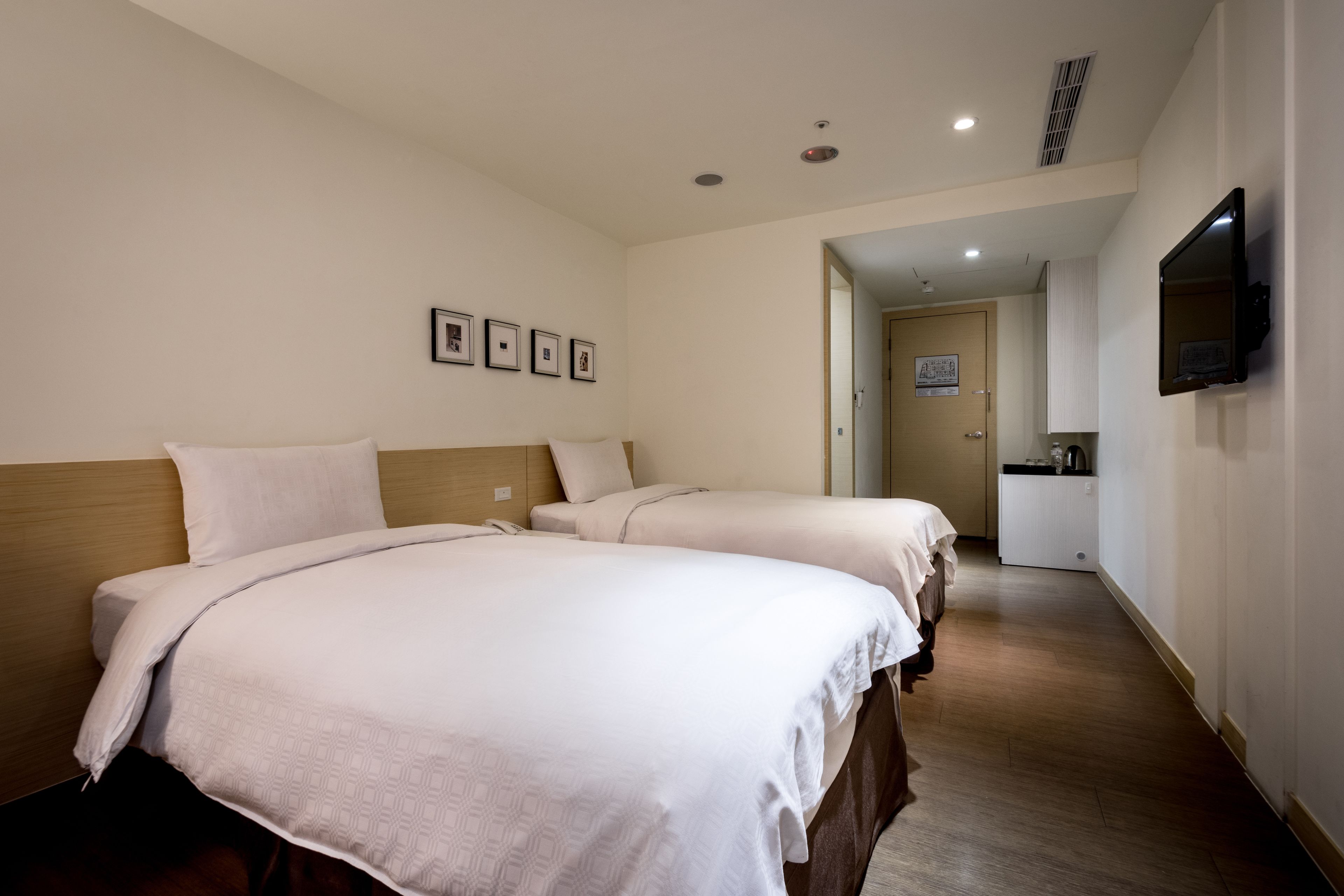 A hotel room with two beds, a TV mounted on the wall, and a door leading to a kitchenette.
