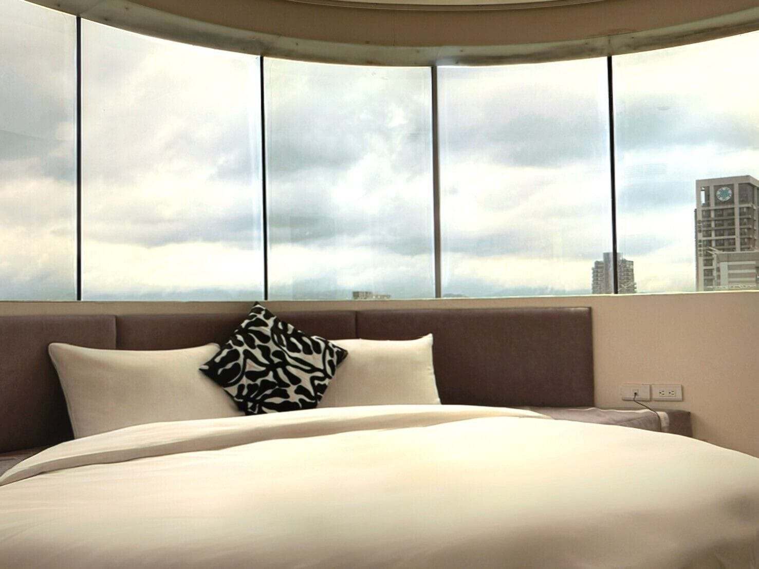 A hotel room with a curved window, a bed with white bedding, and a patterned pillow.