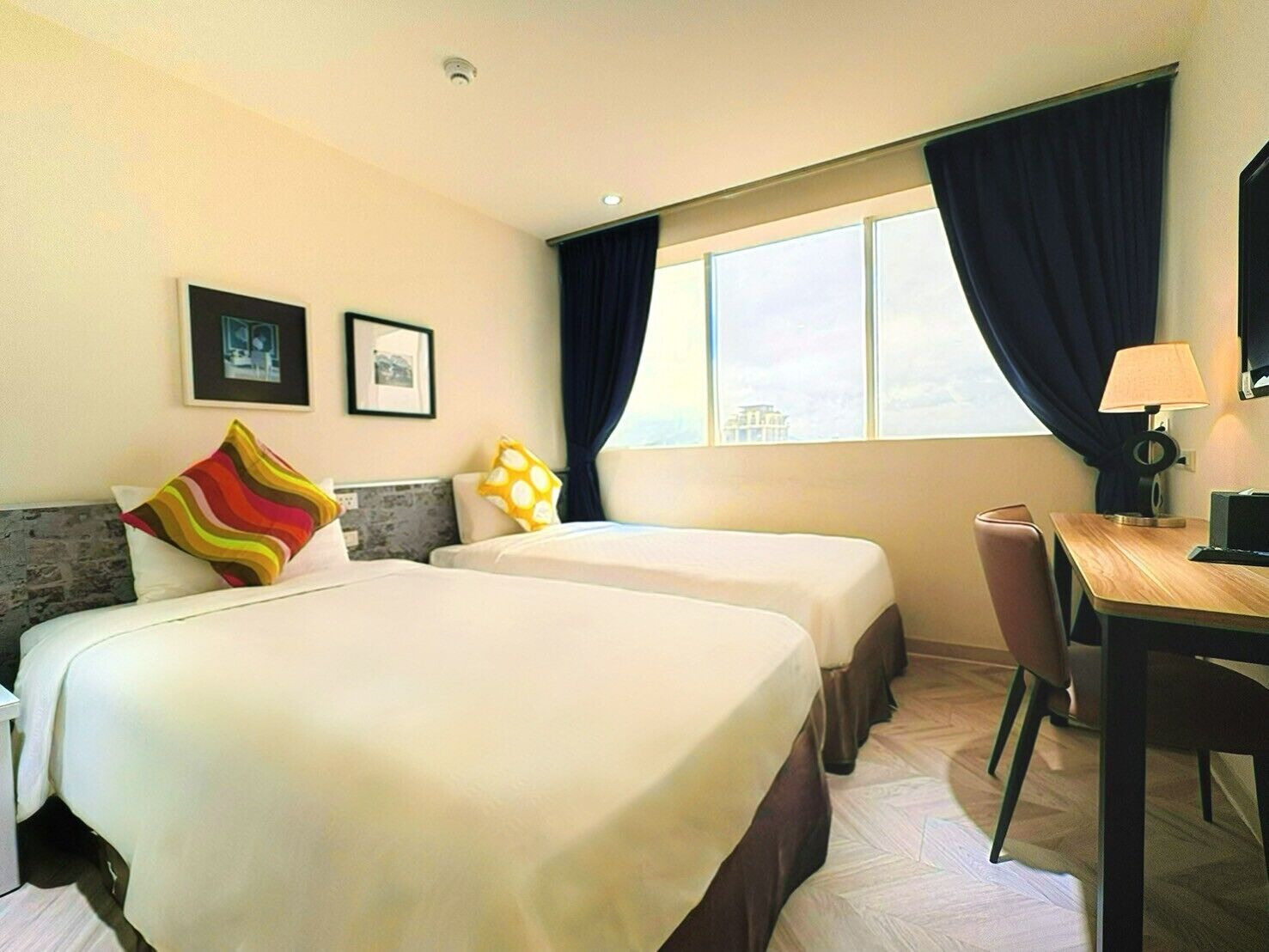 A hotel room with two beds, a desk, a chair, a lamp, and a window with curtains.