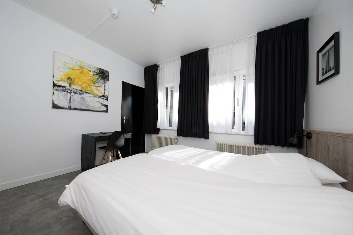 A hotel room with a large bed, a desk, a chair, a radiator, and a painting on the wall.
