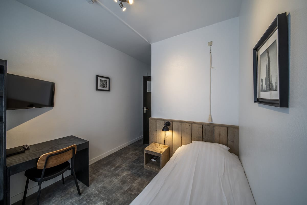 A bedroom with a bed, a desk, and a chair. There is a nightstand with a lamp, a framed picture on the wall, and a door in the background.
