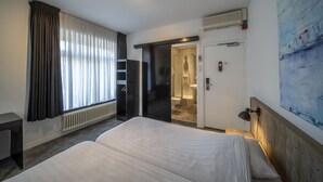 A hotel room with a bed, a desk, a radiator, a shower area, and a painting on the wall. - Hotel St. Lambert (Helmond)