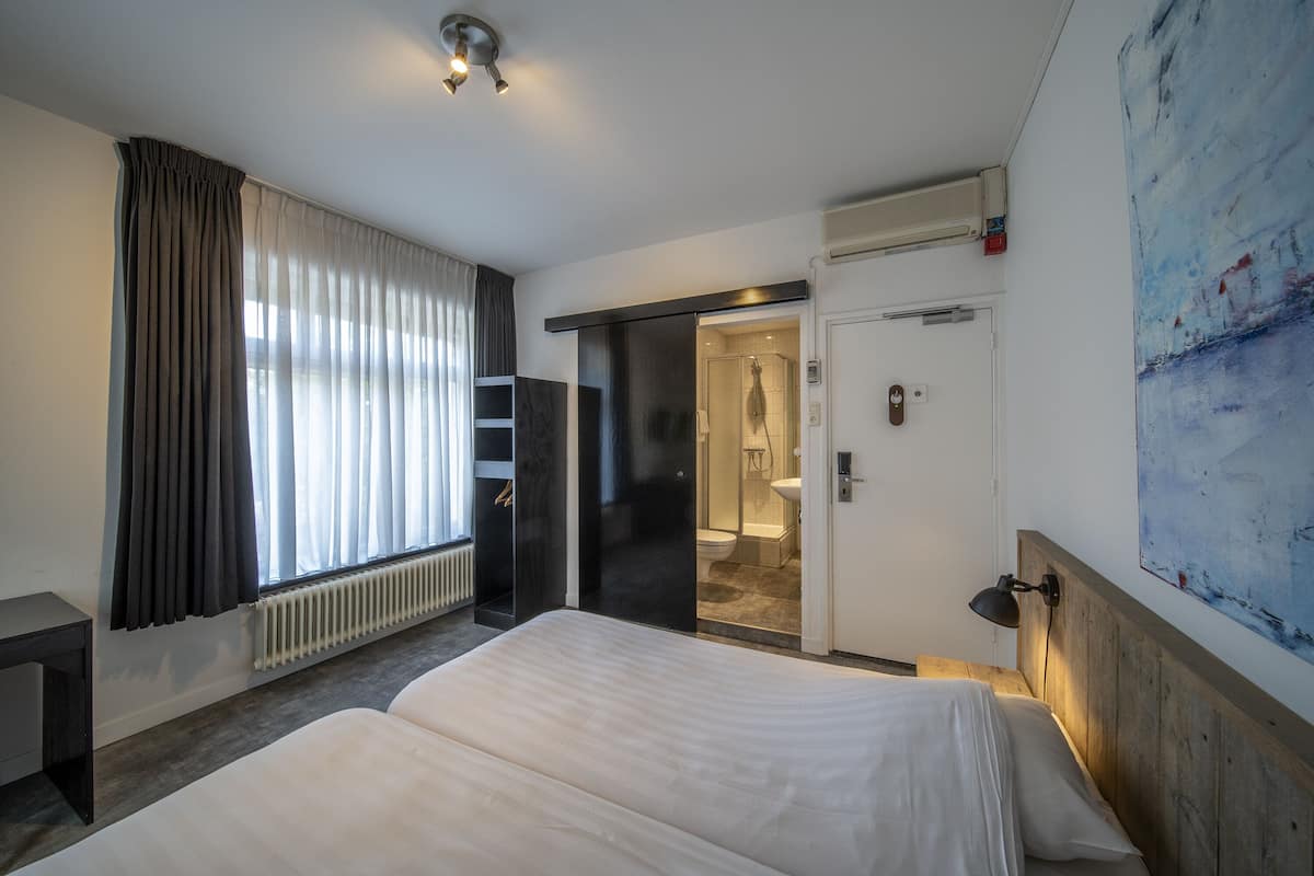 A hotel room with a bed, a desk, a radiator, a shower area, and a painting on the wall.