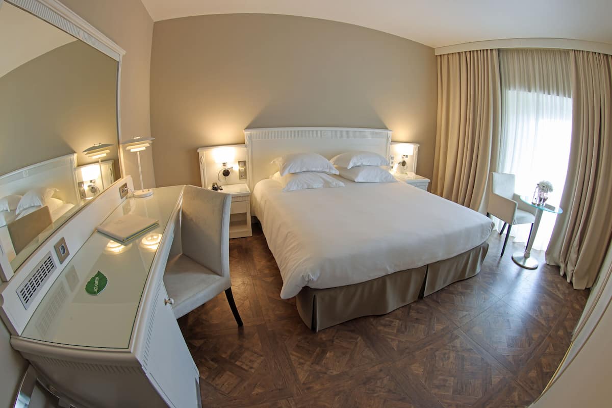 Quarto de hotel com uma cama grande, uma escrivaninha com cadeira, um espelho e uma janela com cortinas.