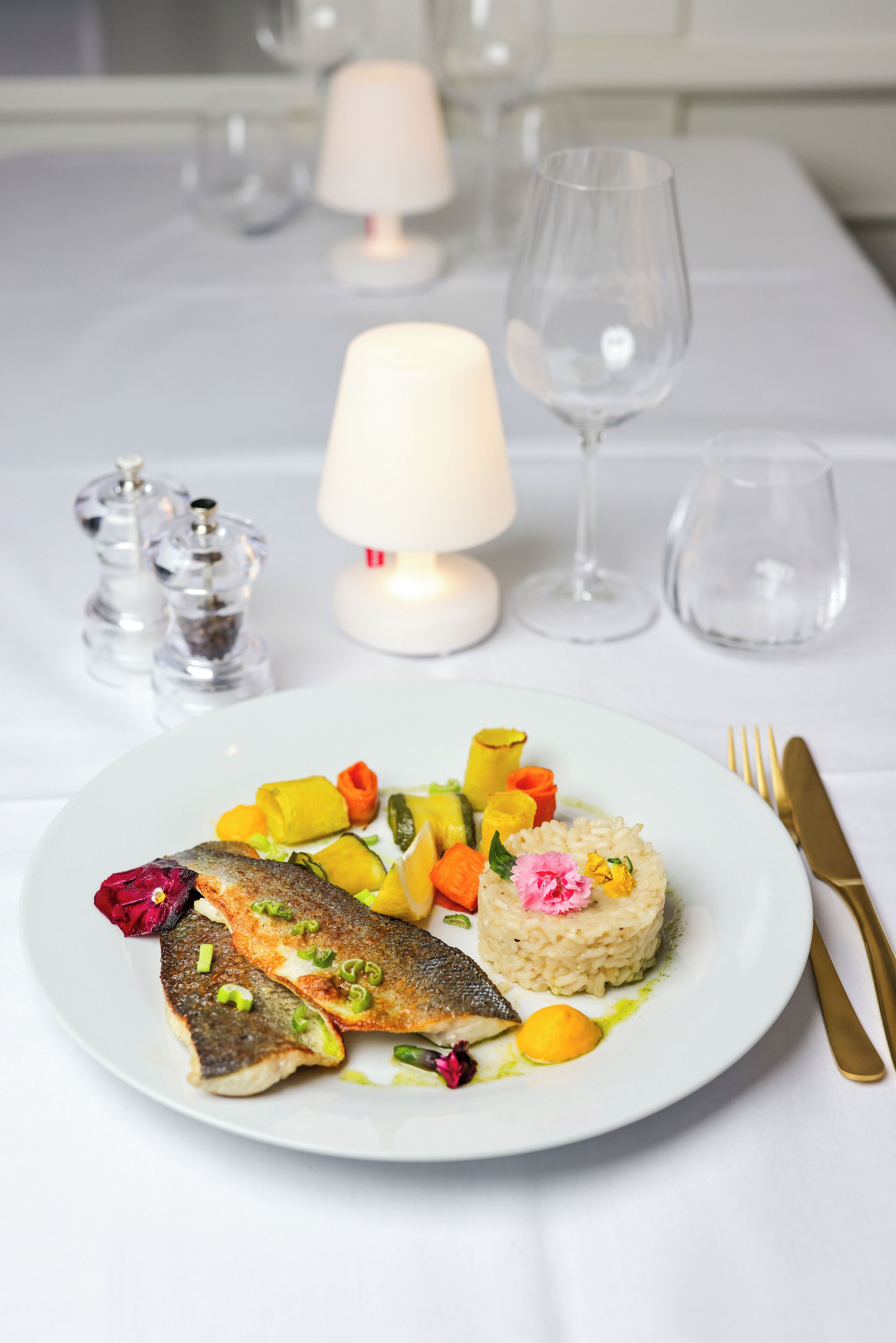 Un plat de menjar amb peix, verdures i arròs, posat sobre un mantell blanc, amb una làmpada i salabros al fons.