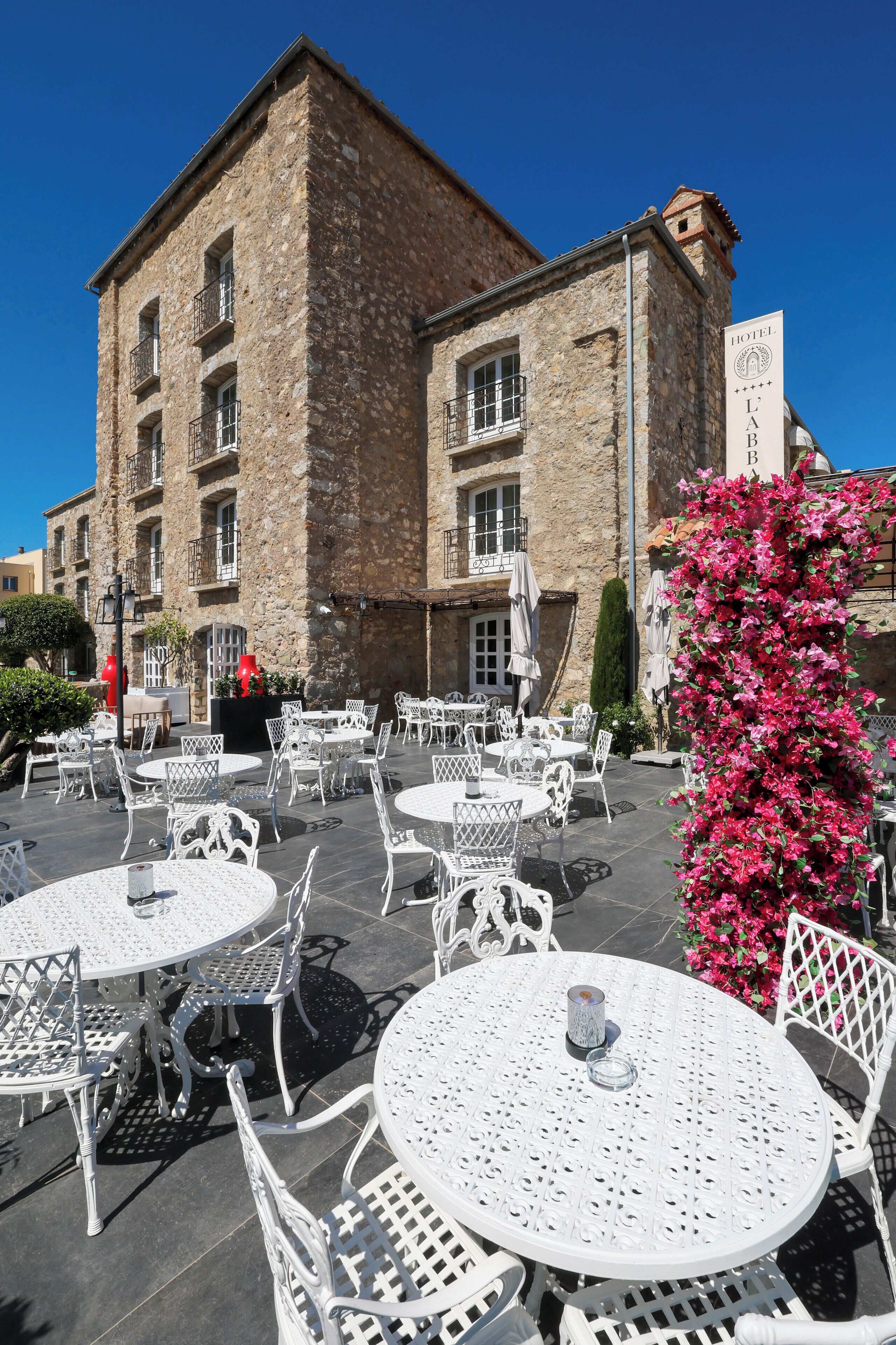 An outdoor patio with white metal tables and chairs, a stone building with balconies, and a sign indicating a hotel.