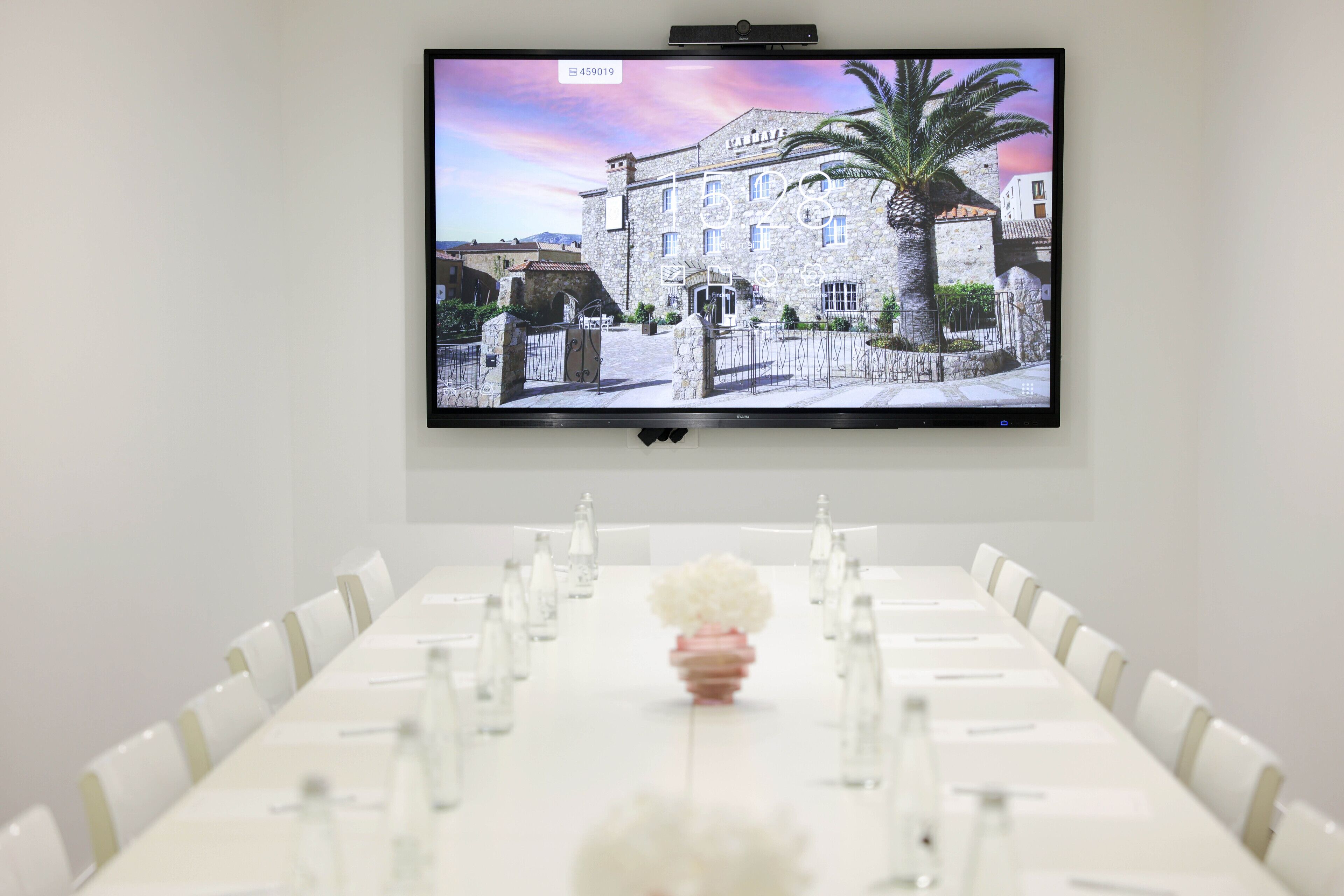 A conference room with a long table set for meetings, a television mounted on the wall displaying a scenic view, and a decorative vase with flowers in the center.