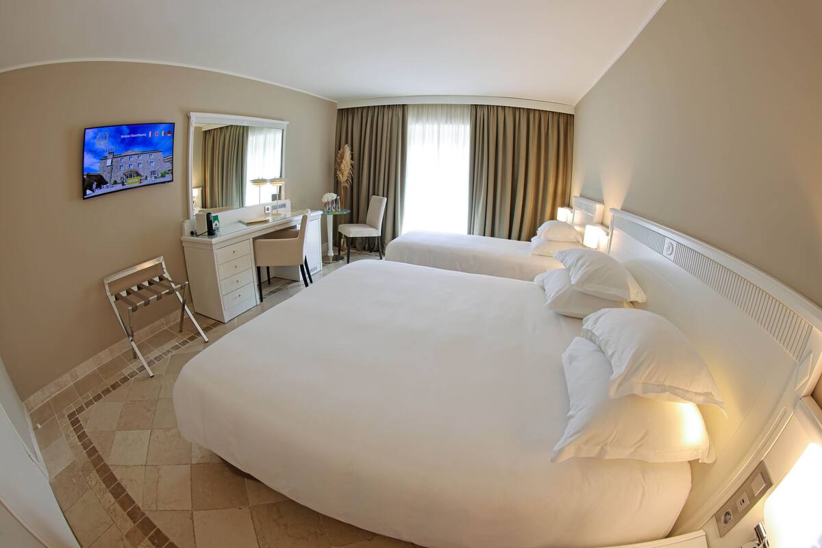 Quarto de hotel com duas camas grandes, uma escrivaninha com cadeira, uma televisão na parede e uma janela com cortinas.