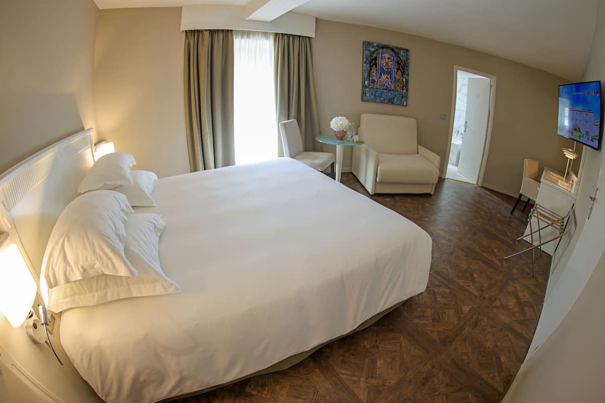 Quarto de hotel com uma cama grande, uma mesinha com um vaso, uma cadeira e uma televisão na parede.