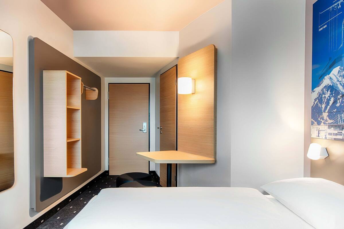 A hotel room with a bed, a mirror, a shelf, and a poster on the wall.
