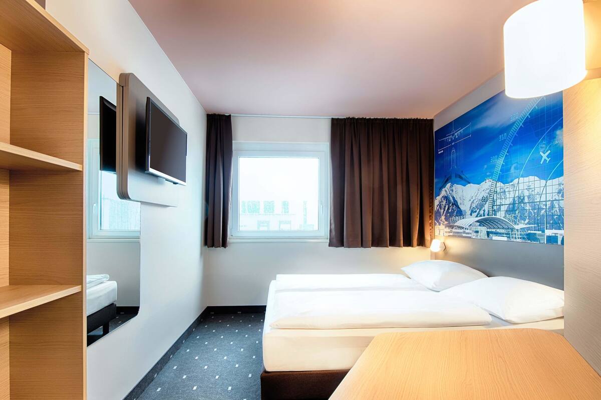 A hotel room with a bed, a TV mounted on the wall, a window with curtains, and a cityscape mural on the wall.