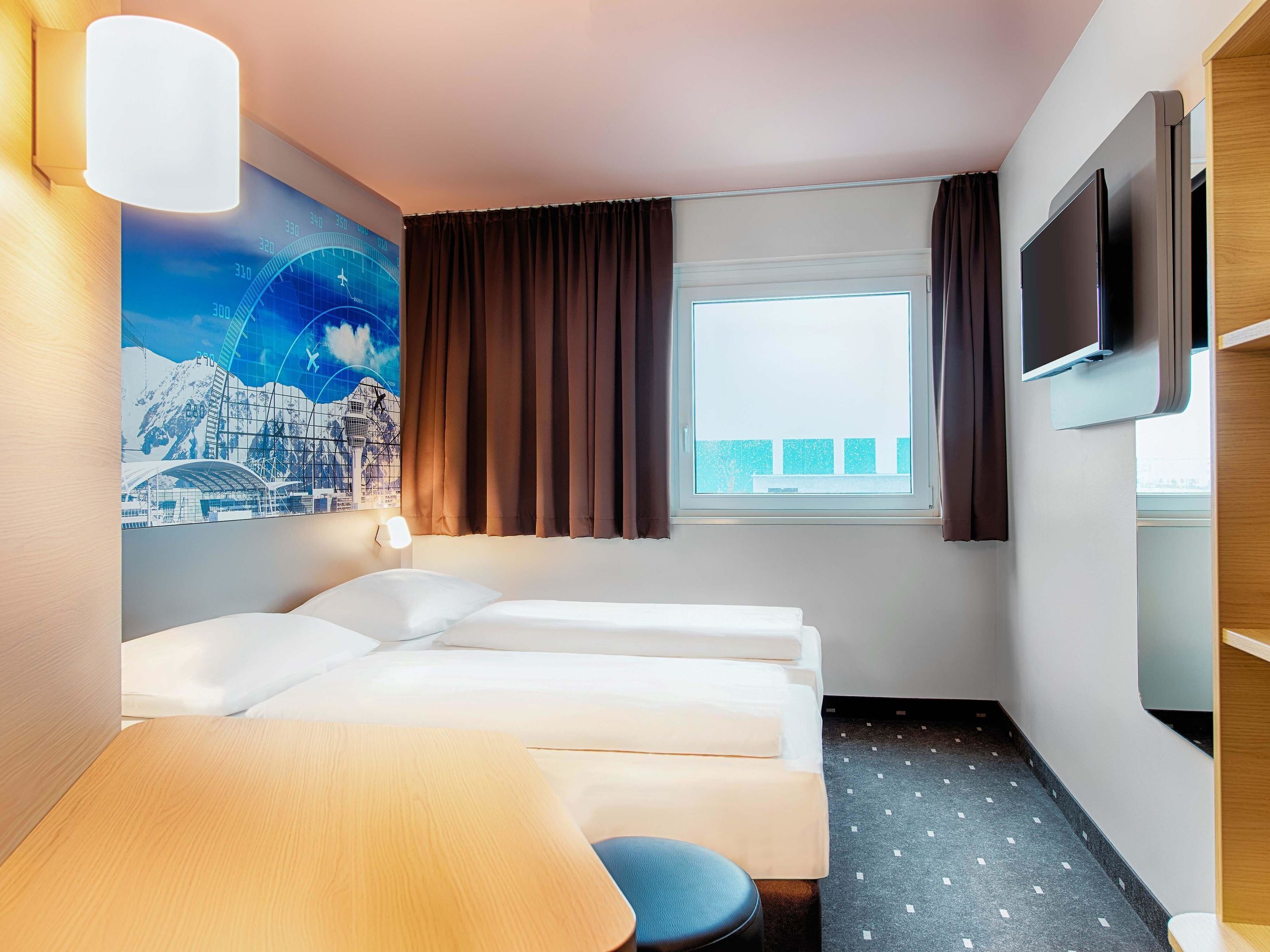 A hotel room with two beds, a desk, a TV, a window with curtains, and a wall with a mountain and cityscape mural.