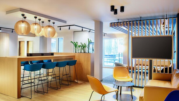 A modern interior with a wooden bar, blue bar stools, a yellow chair, and a large TV screen. - B&B Hotel München-Airport (Hallbergmoos)