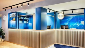 A reception area with a blue color scheme, a bar with shelves of bottles and glasses, and a digital display with the B&B Hotels logo. - B&B Hotel München-Airport (Hallbergmoos)