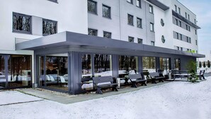 A modern hotel with a covered outdoor seating area and a snow-covered ground. - B&B Hotel München-Airport (Hallbergmoos)