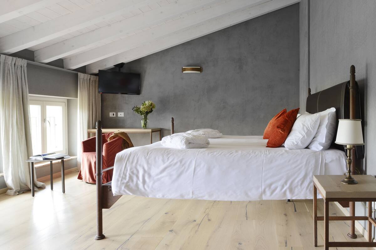A bedroom with a large bed, a bedside table, a lamp, a TV, and a window with curtains.