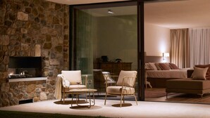 Un intérieur moderne avec un mur en pierre, une cheminée, une porte vitrée donnant sur une chambre, et un coin salon avec deux fauteuils et une table.