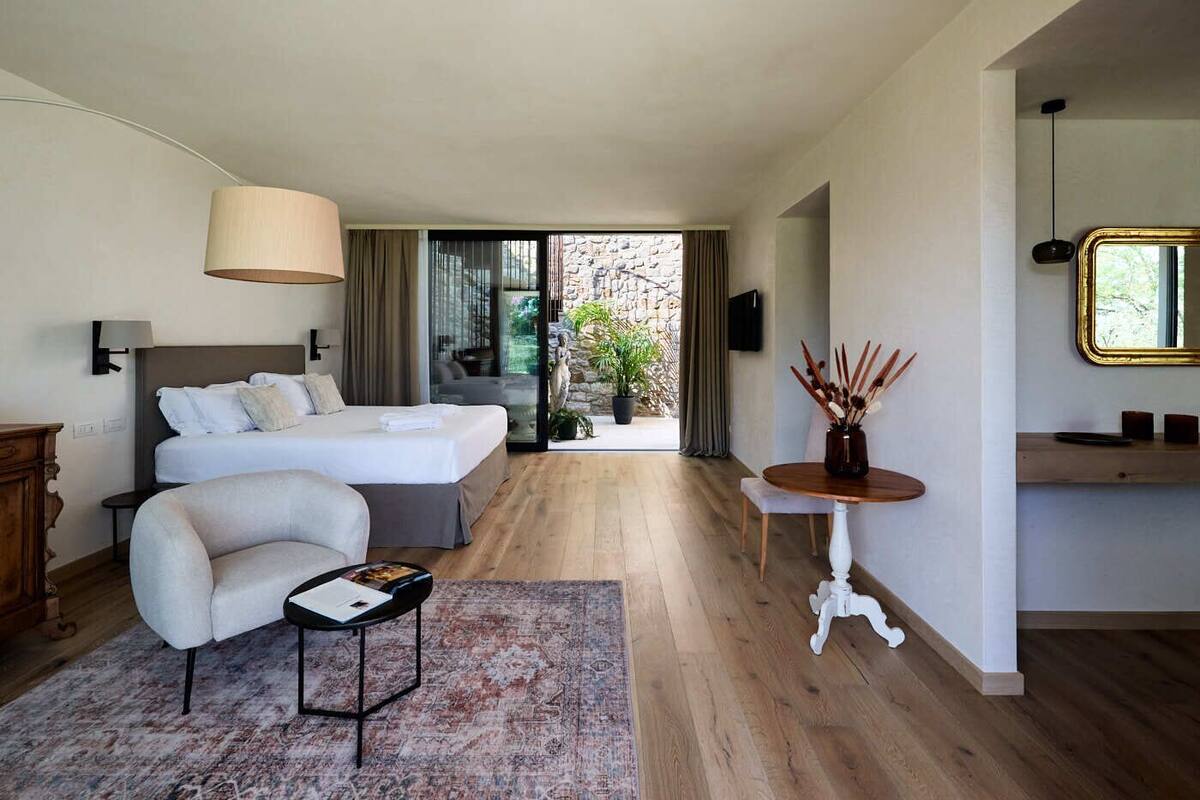 A modern hotel room with a large bed, a seating area with a small table, a large window with a view of the outdoors, and a small side table with decorative items.