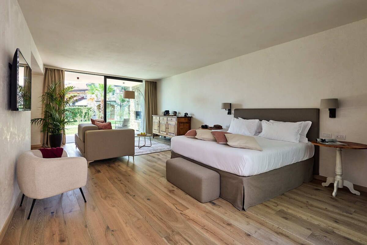 A modern hotel room with a large bed, a sofa, a chair, a small table, and a TV mounted on the wall.