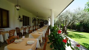Un comedor amplio con mesas dispuestas para varios comensales, una zona de estar techada al aire libre y un jardín con árboles y flores.