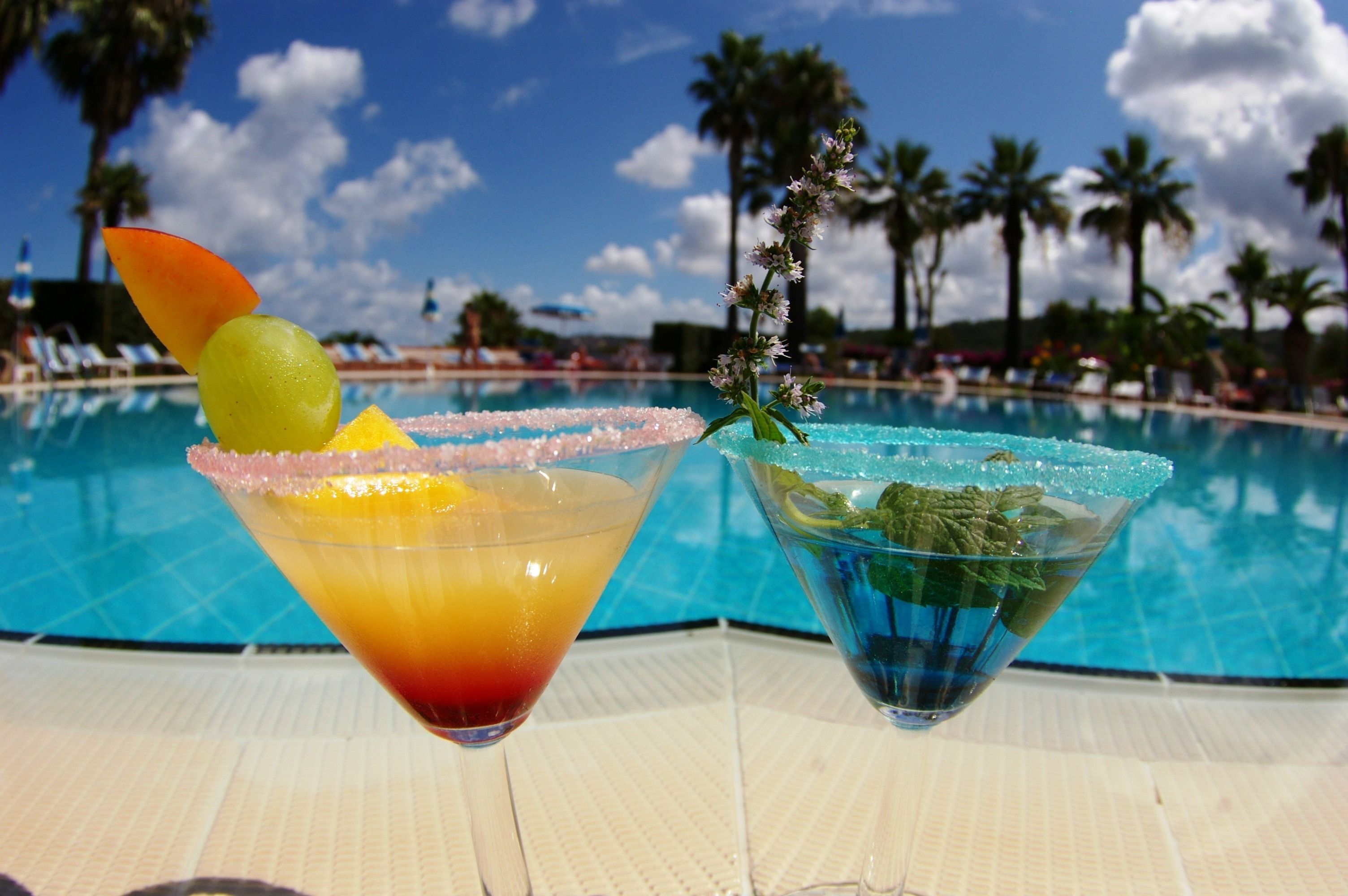 To cocktails på et bord ved en pool med palmer i baggrunden.