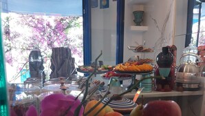 Una mesa de buffet con frutas y verduras diversas, un florero con flores moradas, y una vista de un árbol floreciente de color rosa en el exterior.