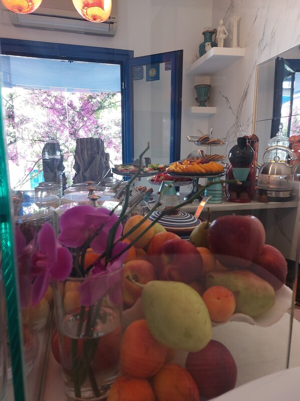 Ein Buffet mit verschiedenen Früchten und Gemüsesorten, eine Vase mit violetten Blumen und der Blick auf einen blühenden rosafarbenen Baum im Freien.