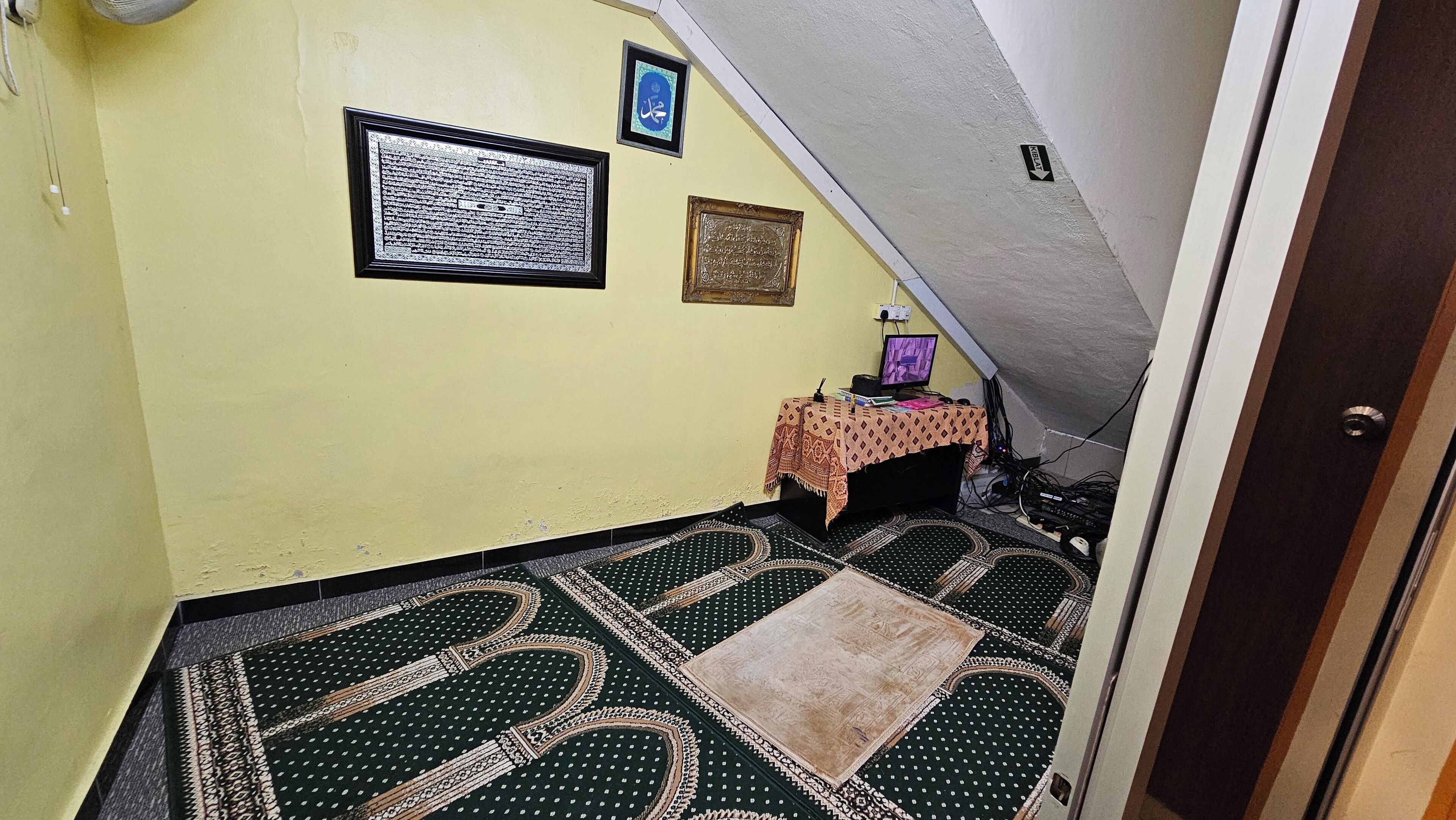 A room with a prayer mat, a small table with a lamp, and framed texts on the wall.