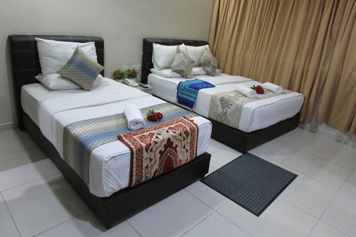 A hotel room with two beds, each with white linens, pillows, and decorative throws. There is a small table with a phone and a vase of flowers.