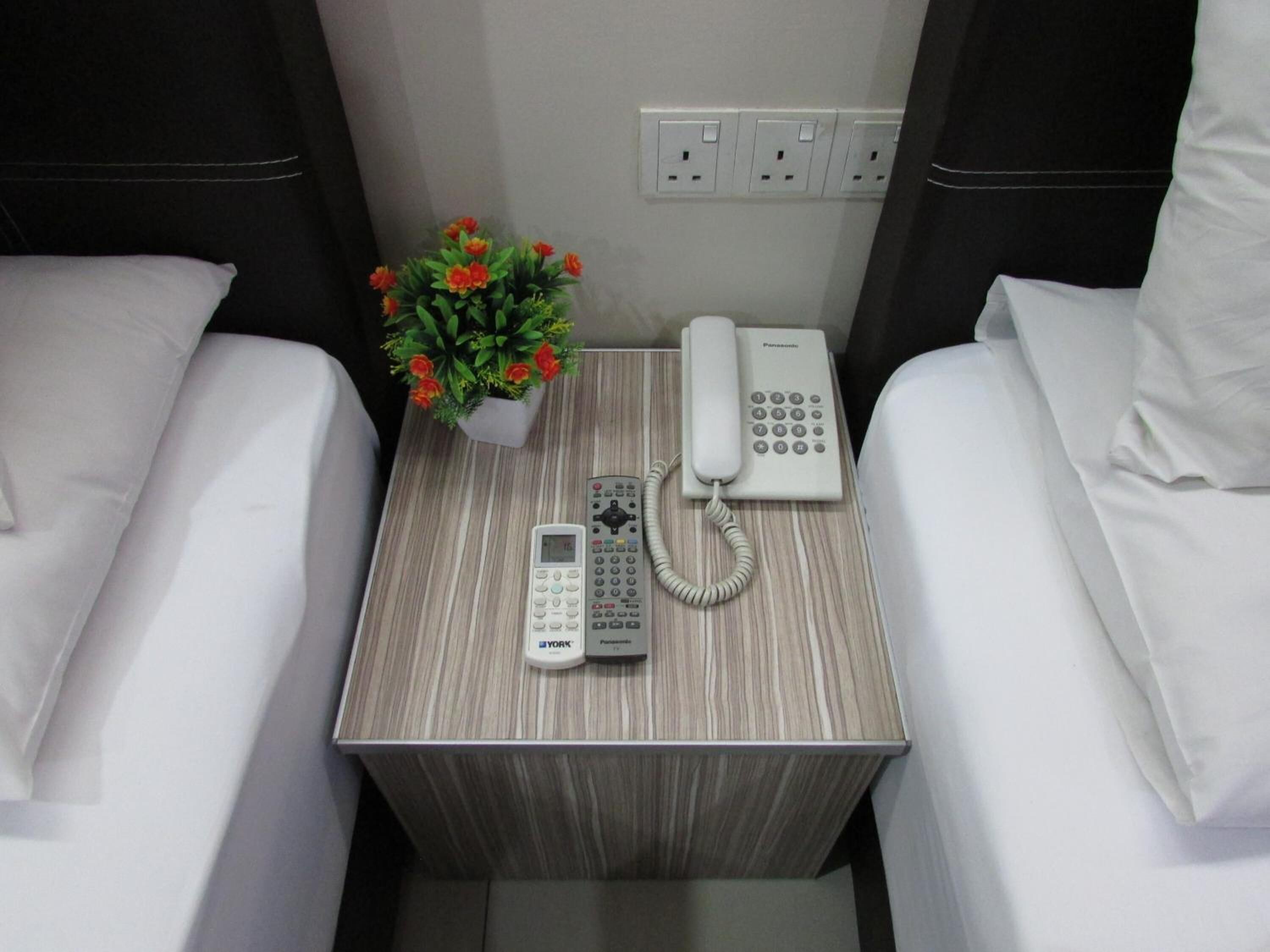 A small bedside table with a Panasonic phone, a York calculator, and a potted plant with red flowers.