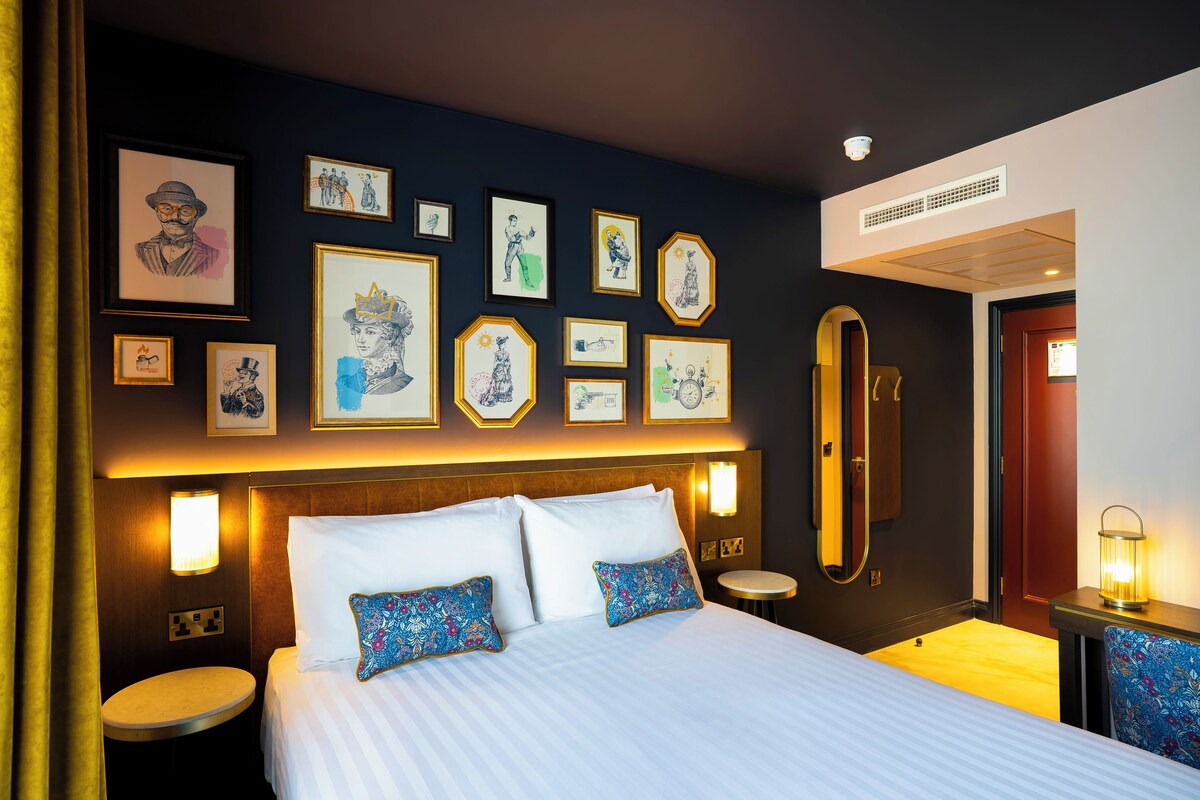 A hotel room with a large bed, bedside tables, a round mirror, and artwork on the walls.