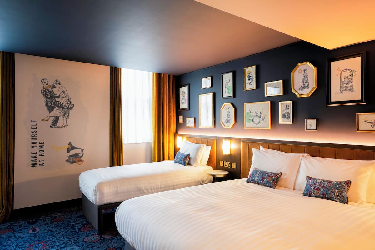 Une chambre d’hôtel avec deux lits, une grande fresque murale et divers tableaux encadrés.