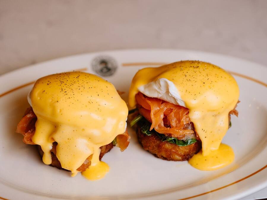 Un plato con dos sándwiches huevos benedictinos cubiertos de salsa holandesa y adornados con un huevo pochado, salmón ahumado y una rodaja de tomate.