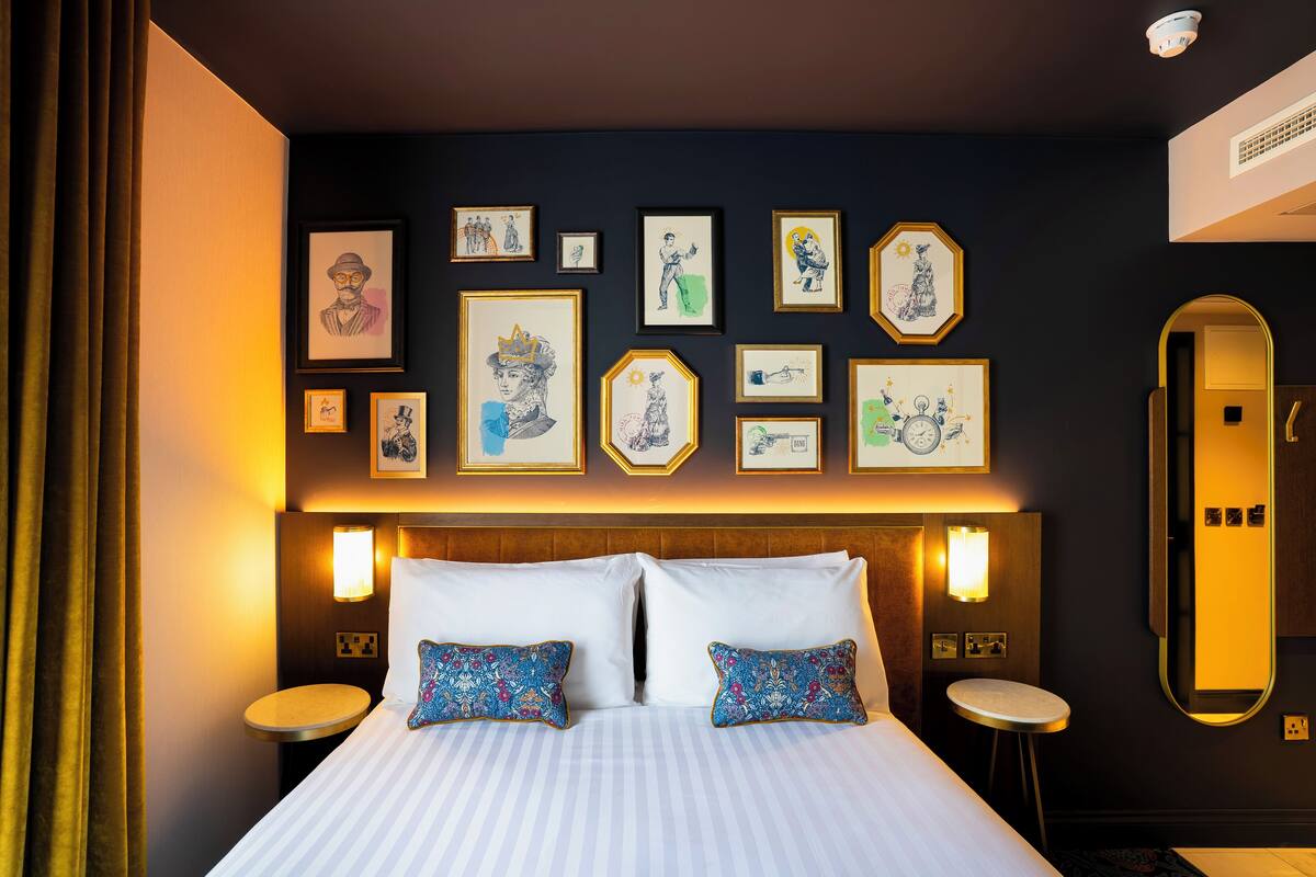 Une chambre d’hôtel avec un grand lit, deux tables de chevet, un miroir fixé au mur et divers tableaux encadrés.