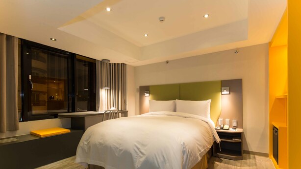 Una habitación de hotel moderna con una cama grande, una mesita de noche con lámpara, una ventana con cortinas y un banco empotrado.