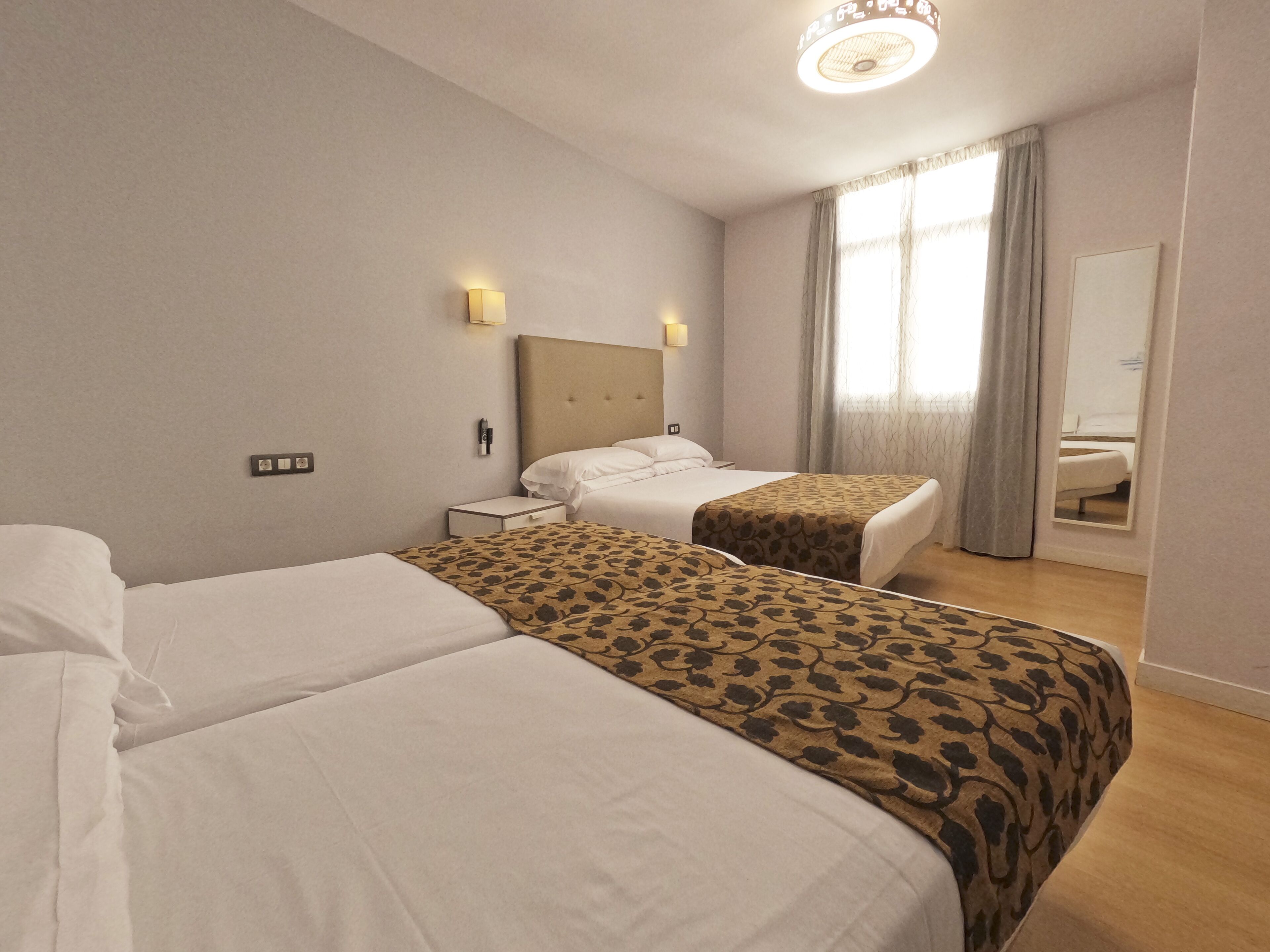 Una camera d'albergo con due letti, un comodino, una lampada a parete, uno specchio e una finestra con tende.